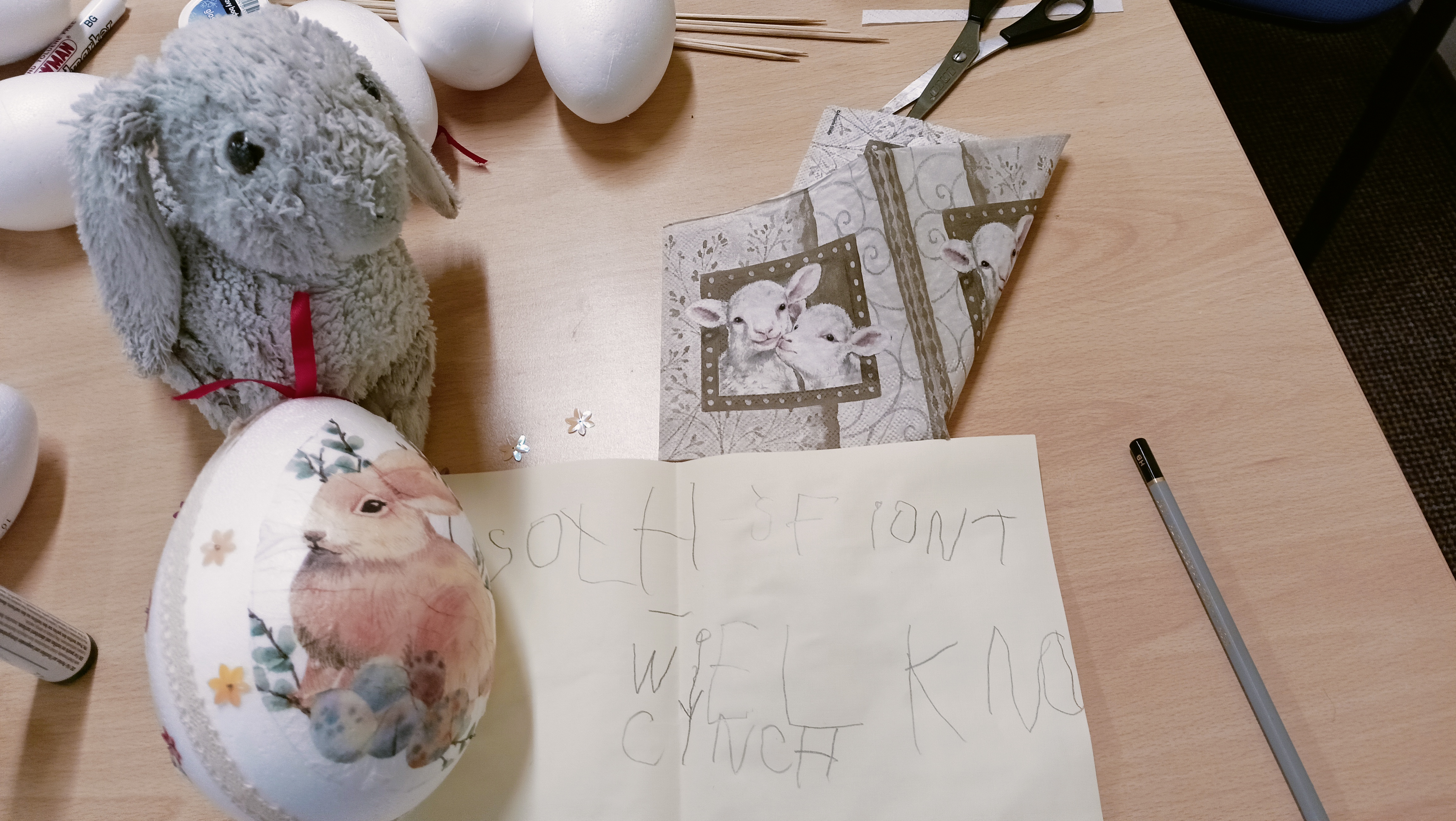 Na stole stoi maskotka królika, przed nim pisanka z rysunkiem królika oraz ręcznie napisane na kartce życzenia wielkanocne
